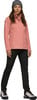 Eine Frau in einer rosa Jacke, schwarzen Hose und gestreiftem Hut steht.