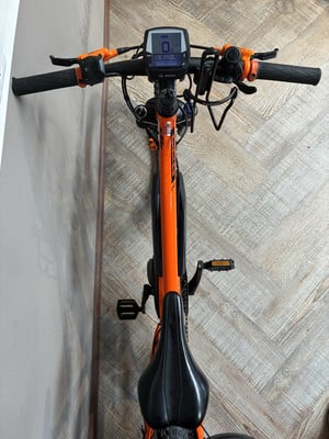 Orange E-Bike mit Display und schwarzem Sattel auf Holzboden. Orange E-Bike mit Display und schwarzem Sattel auf Holzboden.