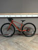 Ein orangefarbenes Gravelbike mit schwarzen Rädern steht auf einem Holzboden vor einer grauen Wand.