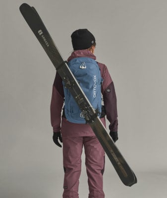 Eine Frau mit Skiern auf dem Rücken und einem blauen Rucksack vor neutralem Hintergrund.