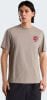 Mann in einem beigen T-Shirt mit Aufdruck auf der Brust und schwarzer Hose.
