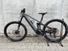 Ein graues E-Mountainbike mit Federung steht auf einem Ständer vor einer Wand.