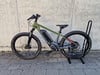 Grünes E-Bike mit schwarzem Ständer auf Kopfsteinpflaster vor einer Wand.