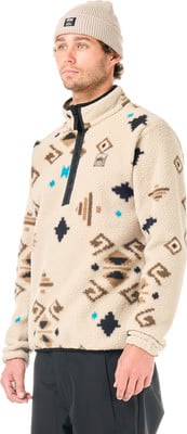 Ein Mann in einem beigefarbenen Fleece-Pullover mit Muster und einer Mütze auf weißem Grund.