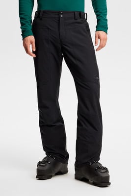 Ein Mann in schwarzer Skihose und grünem Oberteil mit Skischuhen.