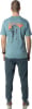 Mann von hinten in blaugrünem T-Shirt mit Bergmotiv und dunkler Hose.