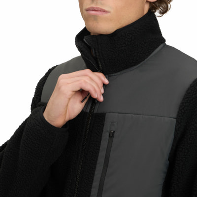 Ein Mann schließt eine schwarze Fleecejacke mit grauen Einsätzen und hohem Kragen.