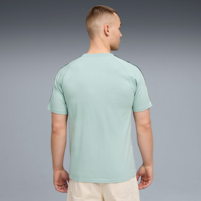 Ein Mann in einem mintgrünen T-Shirt mit Streifen an den Ärmeln und einer beige Hose.