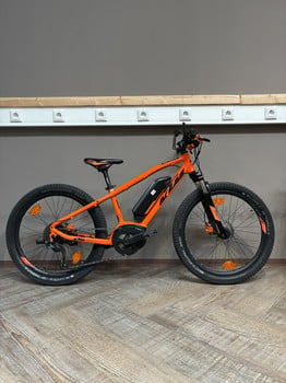 Ein orangefarbenes KTM E-Bike steht auf einem Holzboden vor einer grauen Wand mit weißen Steckdosen.