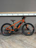 Ein orangefarbenes KTM E-Bike steht auf einem Holzboden vor einer grauen Wand mit weißen Steckdosen.