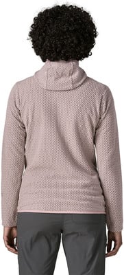 Rückansicht einer Frau mit lockigem Haar in einem rosa Pullover und einer grauen Hose.