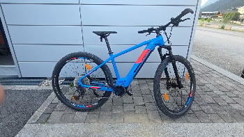 Ein blaues E-Bike mit orangefarbenen Akzenten, das vor einem Gebäude geparkt ist.