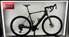 Ein graues E-Bike mit gelber Gabel steht auf einem Ständer vor einer weißen Wand.