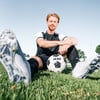 Ein Fußballer sitzt mit einem Ball und Fußballschuhen auf dem Rasen.