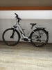 Modernes silbernes E-Bike mit Schutzblechen und Gepäckträger, drinnen abgestellt.