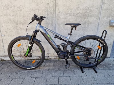 Ein graues E-Bike mit schwarzen Details steht auf einem Ständer vor einer Wand.