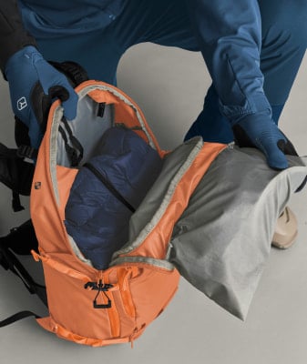 Eine Person packt eine blaue Jacke und eine graue Plane in einen orangefarbenen Rucksack. Eine Person packt eine blaue Jacke und eine graue Plane in einen orangefarbenen Rucksack.