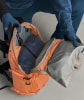 Eine Person packt eine blaue Jacke und eine graue Plane in einen orangefarbenen Rucksack.