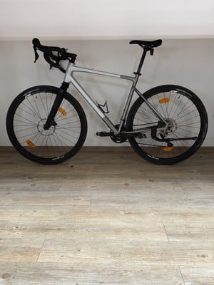 Ein graues Gravelbike mit schwarzen Rädern und Lenker steht auf einem Holzboden. Ein graues Gravelbike mit schwarzen Rädern und Lenker steht auf einem Holzboden.