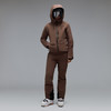 Ein Model in brauner Skijacke, Hose und Skischuhen posiert vor grauem Hintergrund.