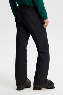 Ein Mann in schwarzer Skihose und grünem Pullover trägt Skischuhe.
