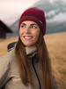 Eine Frau in einer Strickmütze und Jacke lächelt mit Bergen im Hintergrund.