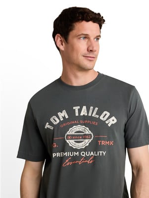 Férfi sötét pólóban „TOM TAILOR ORIGINAL SUPPLIES since 1962” felirattal. Férfi sötét pólóban „TOM TAILOR ORIGINAL SUPPLIES since 1962” felirattal.