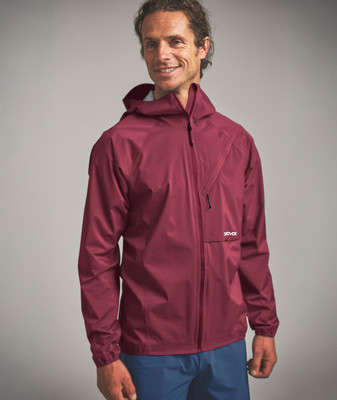 Ein Mann in einer dunkelroten Kapuzenjacke und blauen Hosen lächelt.