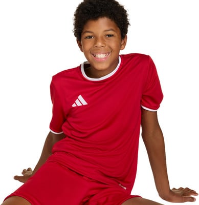 Ein Junge in einem roten Fußballtrikot mit weißem Besatz und Adidas-Logo sitzt. Ein Junge in einem roten Fußballtrikot mit weißem Besatz und Adidas-Logo sitzt.