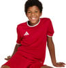 Ein Junge in einem roten Fußballtrikot mit weißem Besatz und Adidas-Logo sitzt.