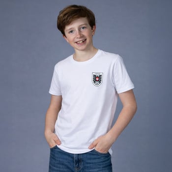 Ein Junge im weißen T-Shirt mit Logo auf der Brust und Jeans steht vor grauem Hintergrund.