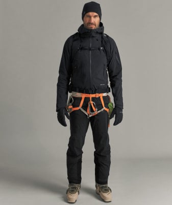 Ein Mann in Skibekleidung mit Klettergurt und Ausrüstung steht vor neutralem Hintergrund.