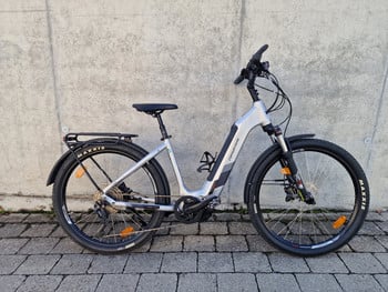 Ein silbernes E-Bike mit schwarzen Akzenten und Schutzblechen steht vor einer Wand.