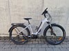 Ein silbernes E-Bike mit schwarzen Akzenten und Schutzblechen steht vor einer Wand.
