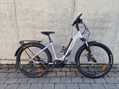 Ein silbernes E-Bike mit Schutzblechen und Gepäckträger steht an einer Wand.