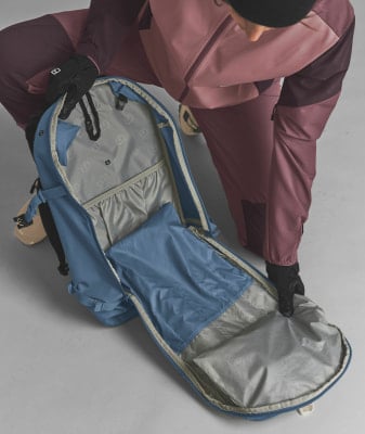 Eine Person in Handschuhen betrachtet das Innere eines blauen Rucksacks mit verschiedenen Fächern.